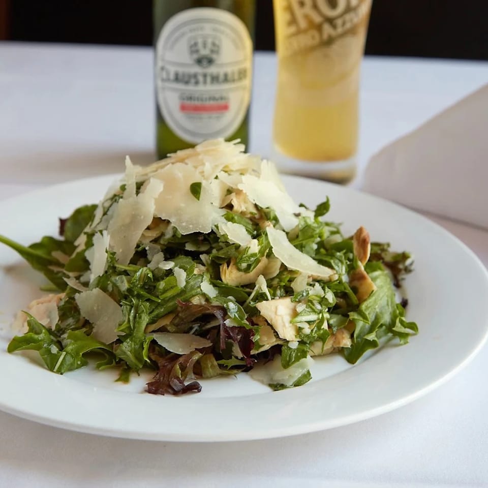 Mixed green salad topped with shaved Parmesan and sliced chicken, served on a white plate with a beer and lemonade.