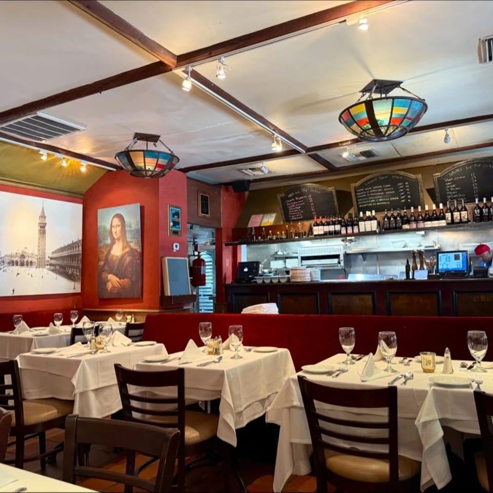 Elegant Italian restaurant dining room with white tablecloths, wine bottles, colorful stained-glass lights, and Venetian artwork.