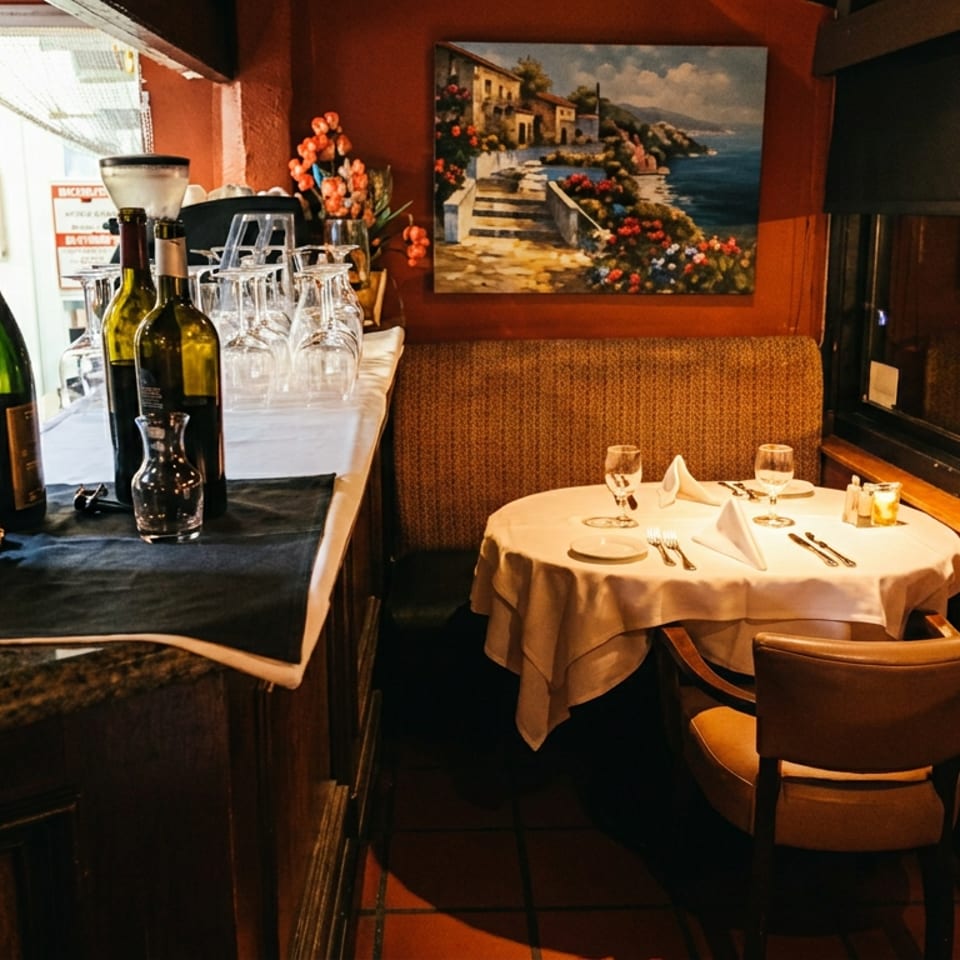 Cozy restaurant interior with a set table, wine bottles, and a painting of a coastal town.